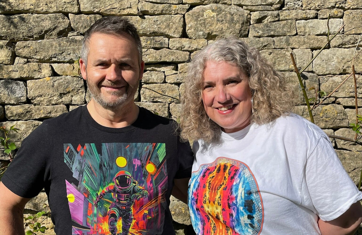 Two people wearing colorful t-shirts with space-themed designs standing in front of a stone wall.